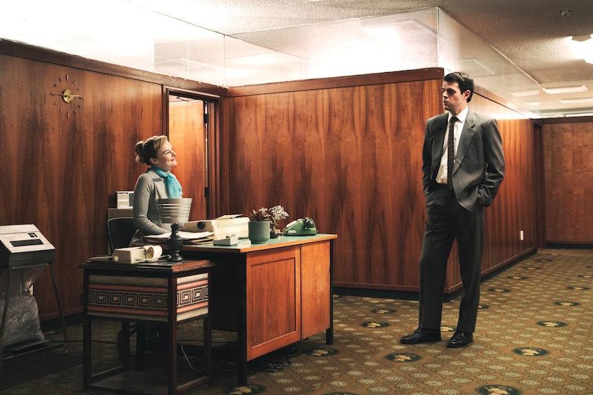 Abby Howells sits behind a desk in a very stylised 60s office, Paul Williams stands in a suit talking to her