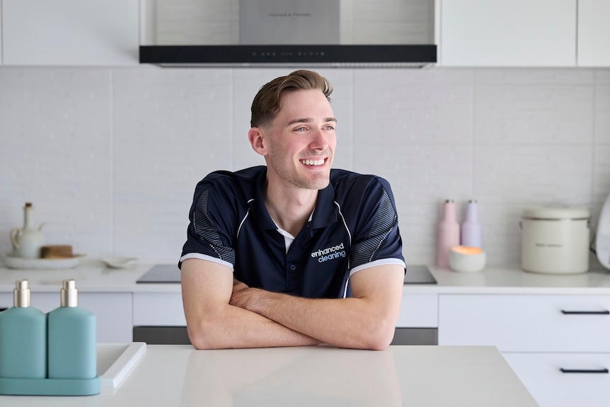 A smiling young man looking off camera leans on his elbows in a clean white kitchen.