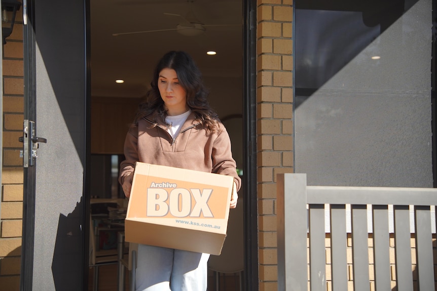 A woman walks through an open door holding a moving box.