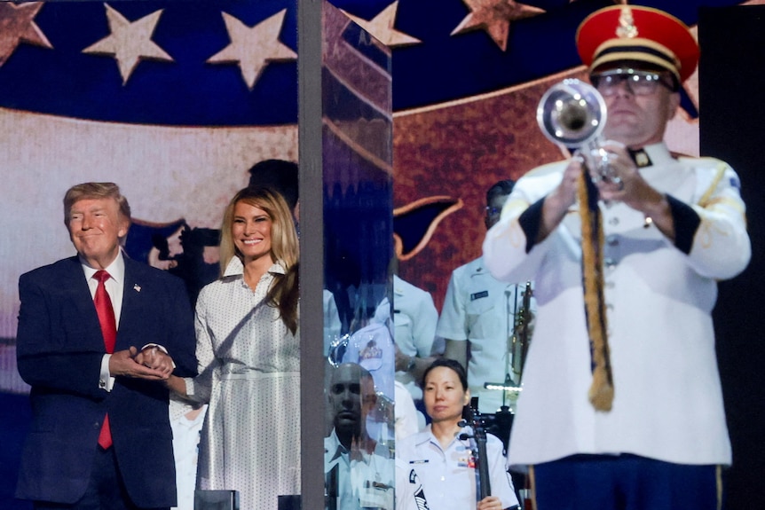 Donald and Melania Trump smile on stage as trumpet player stands in front