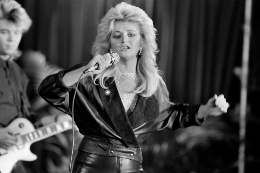 A blonde woman holds a microphone and wears a leather top while erforming on stage.