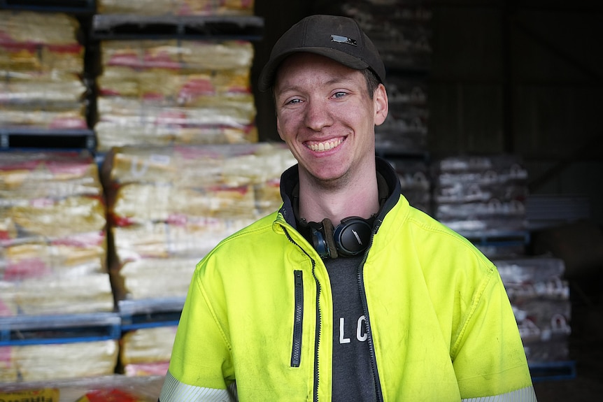 A young man in a baseball cap, with dirt on his face, in front of stacks of firewood.