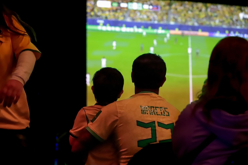 A young boy sits on father's lap amid seats football fans watching a large screening of the world cup