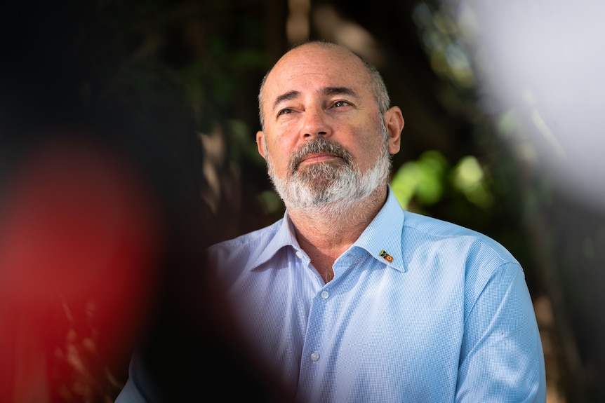 A bald man with a grey beard and a moustache in a light blue shirt looks to the right