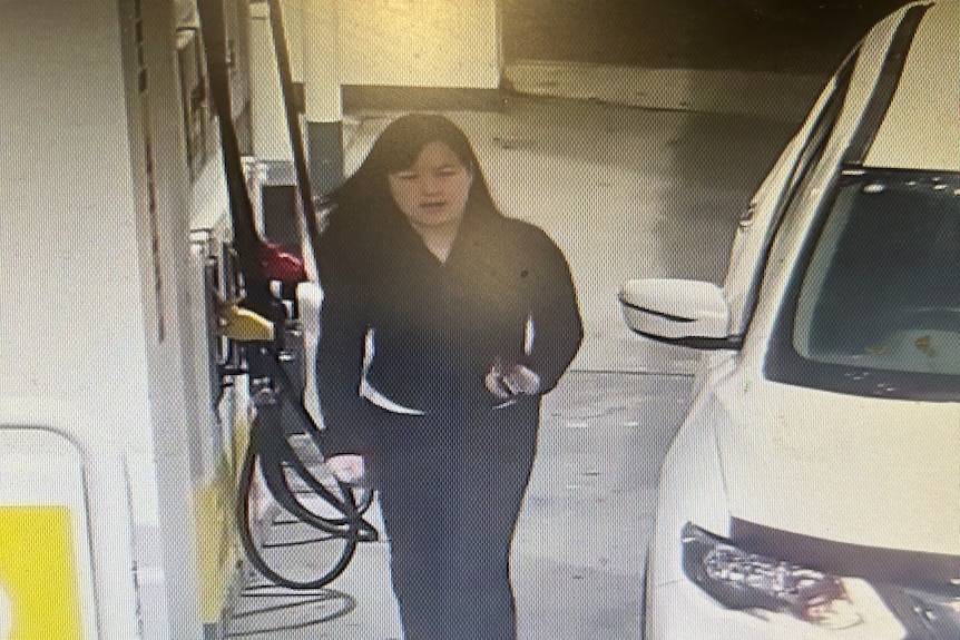 An image from CCTV footage of a dark haired woman in a black top and pants walking between a petrol bowser and a white car.