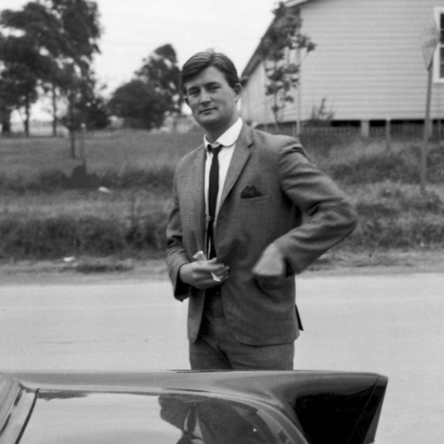 A black and white photo of a man in a light coloured suit with a black tie standing near a car with his hand in his pocket.