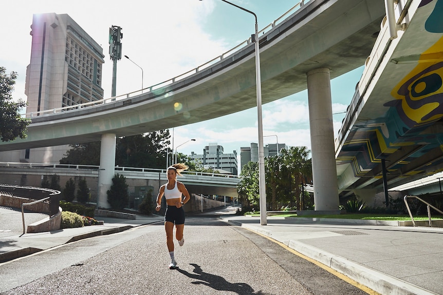 A woman with long hair runs under an overpass in a city.