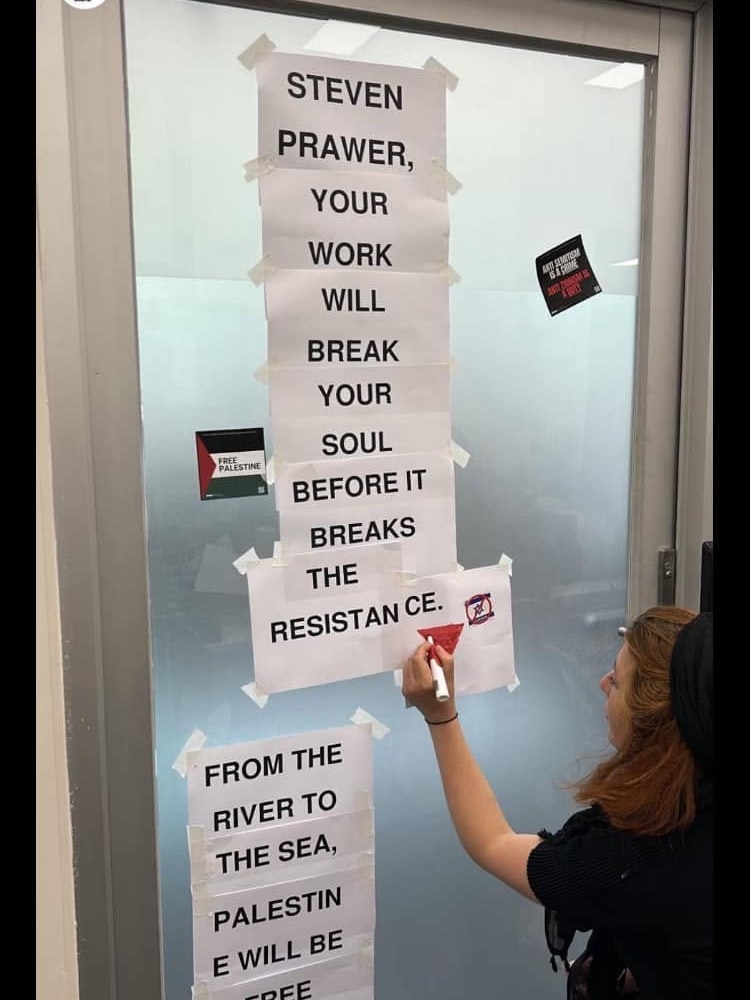 A woman draws on one of a series of a pieces of paper stuck on a door saying "Steven Prawer, your work will break your soul".