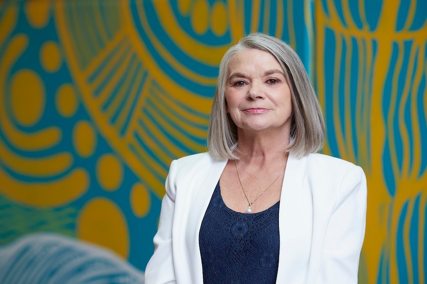A woman with silver hair wears a white blazer and stands in front of a wall painted with an intricate design.