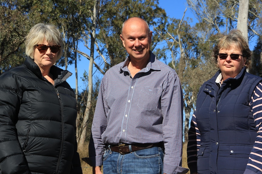 Two women and a man stand in front of some bushland,