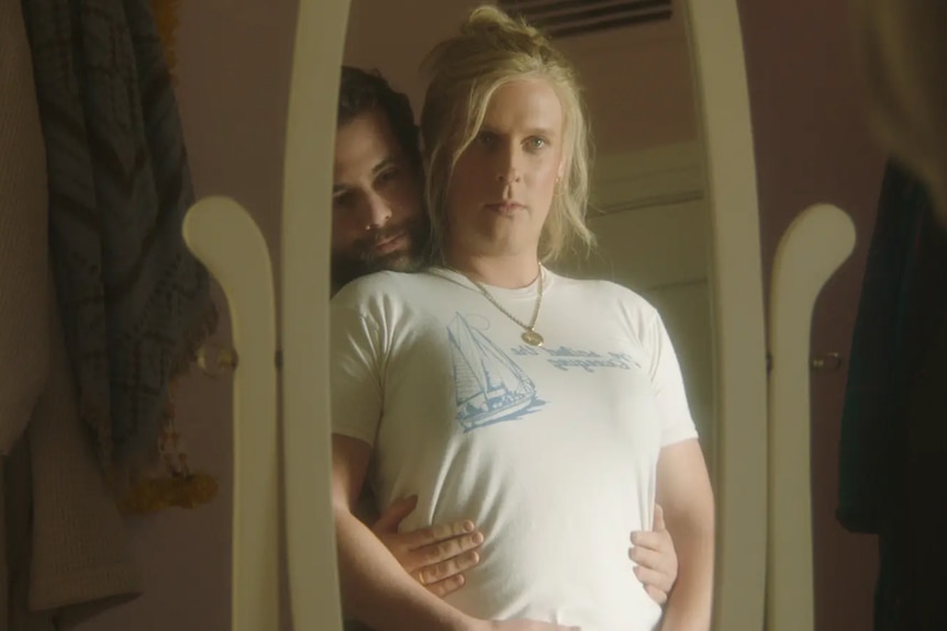 A blonde woman in a white shirt looks at herself in the mirror. A man has his head on her shoulder and hands on her waist