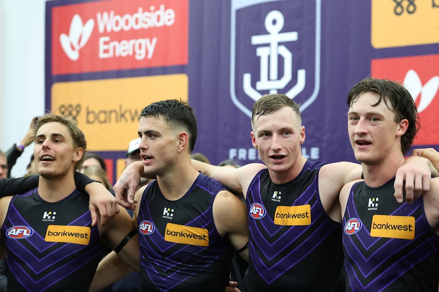 Four Fremantle AFL players (including three talls) stand in the rooms singing the club song after a win.