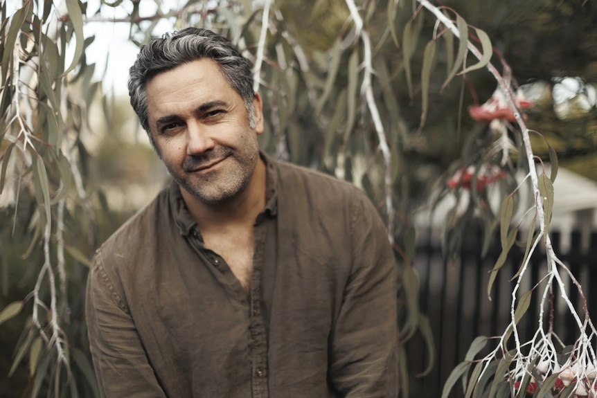 James Henry wears a brown shirt standing among eucalyptus branches with gum blossoms, outdoors in soft natural light.