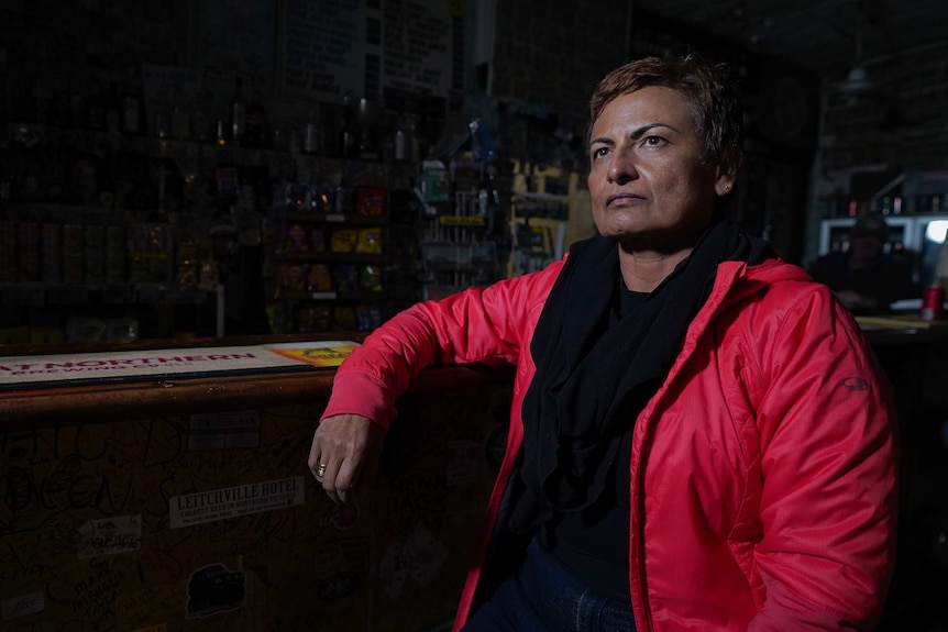 Sitting on a bar stool and leaning on the countertop, a woman in a red jacket wears a tense facial expression.