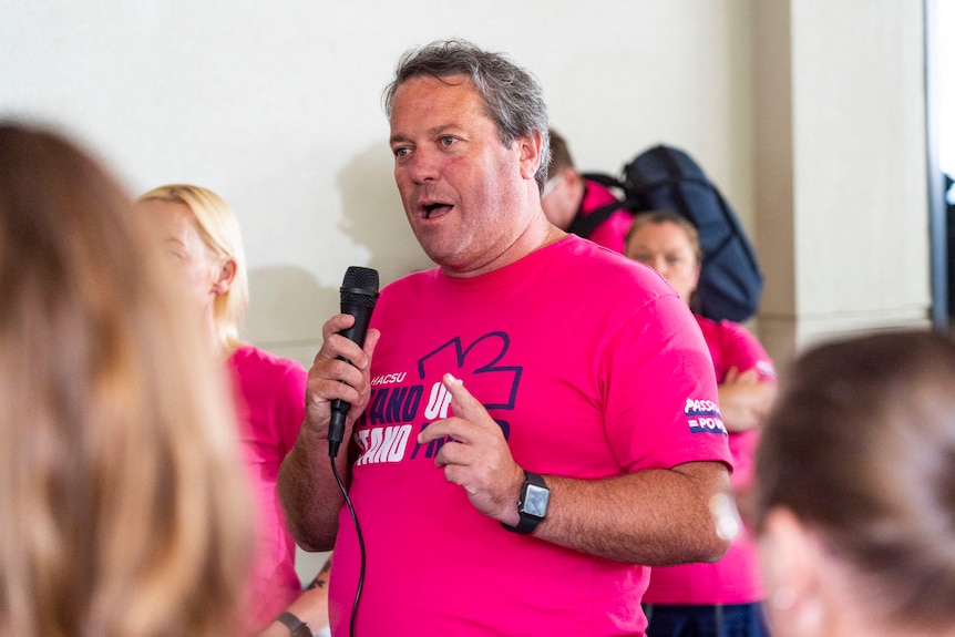 A man with grey hair in a pink shirt speaks on a microphone in front of a crowd.