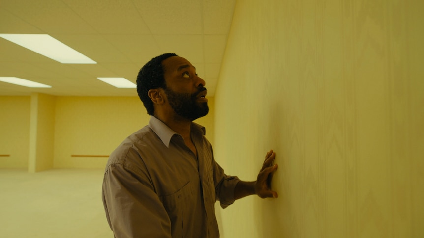 A Black mans puts a hand on the wall and looks up at the ceiling in a yellowed room