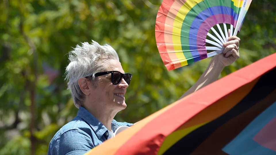 Johnny Knoxville with short white hair and sunglasses holds a rainbow fan and flag, outdoors with trees in the background.