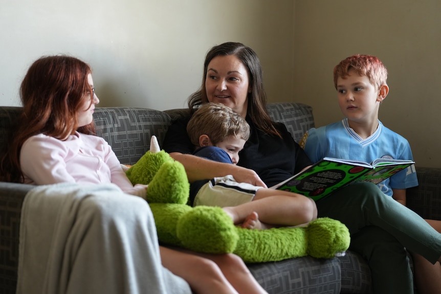A woman sitting on couch with three kids, a young girl with auburn hair, a young boy in her arms and another boy next to her