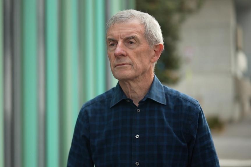 A man with white hair in a blue checked shirt stares to the right of the camera.
