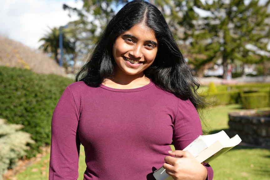 A woman with black hear wearing a maroon shirt holds books.