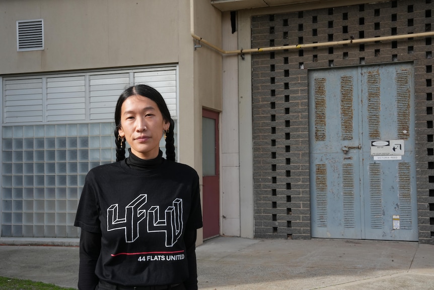 A young Asian woman in pigtails and branded T-shirt looks at camera outside high rise apartment