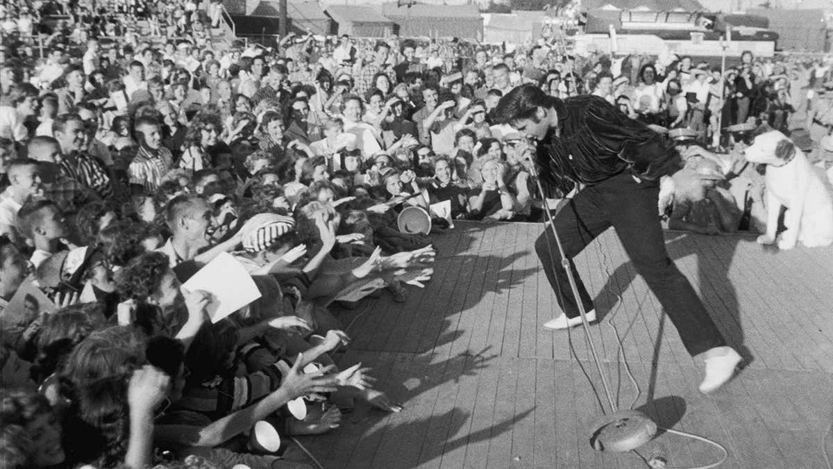 Elvis Presley performs outdoors on a small stage before a crowd.