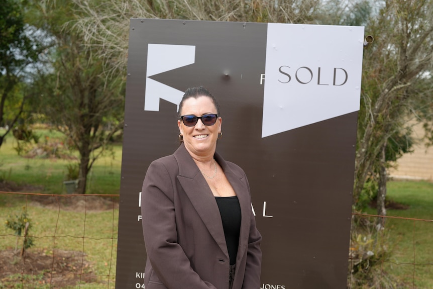 A woman stands smiling in front of a sign with a sold sticker on it.
