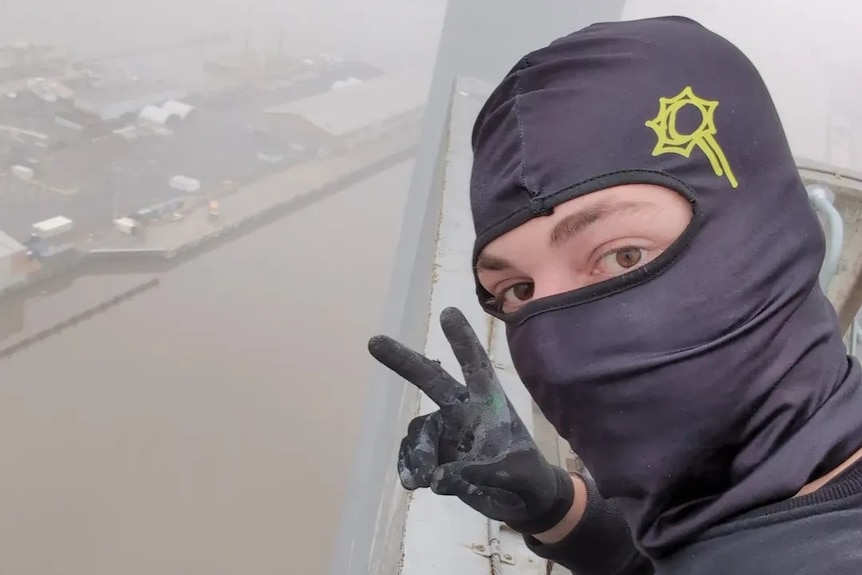 A person with brown eyes in a face covering holds up two fingers while sitting on the edge of a building above a river.