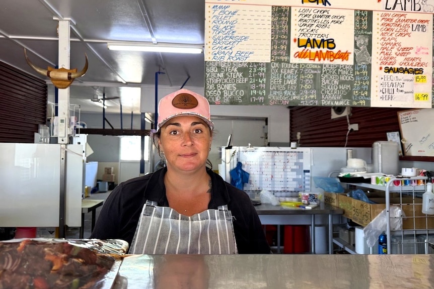 Female butcher in cap and apron standing behind the counter of her store with kebabs in front of her and beef/lamb sign.
