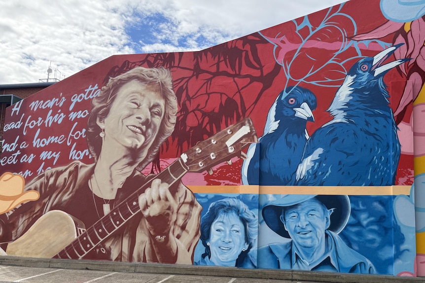 A blue sky sits above a painted wall mural showing a woman smiling and playing the guitar