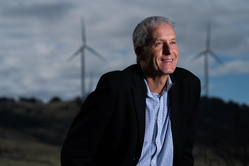 A smiling, grey-haired man in a field with wind turbines.