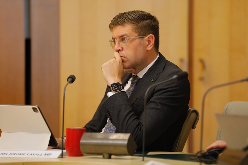 A dark-haired man in a suit sits at a long table in a parliamentary chamber.