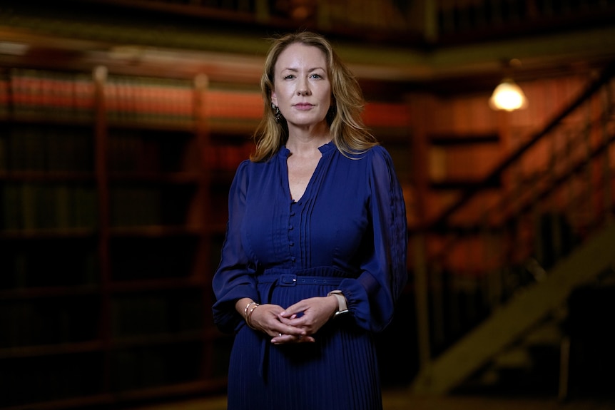 A woman in a blue dress stands holding her hands together as she stands in a darkened room with a large bookcase.
