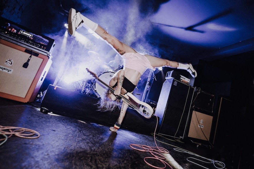 A woman on stage doing the splits in a one-handed handstand while holding a bass guitar.