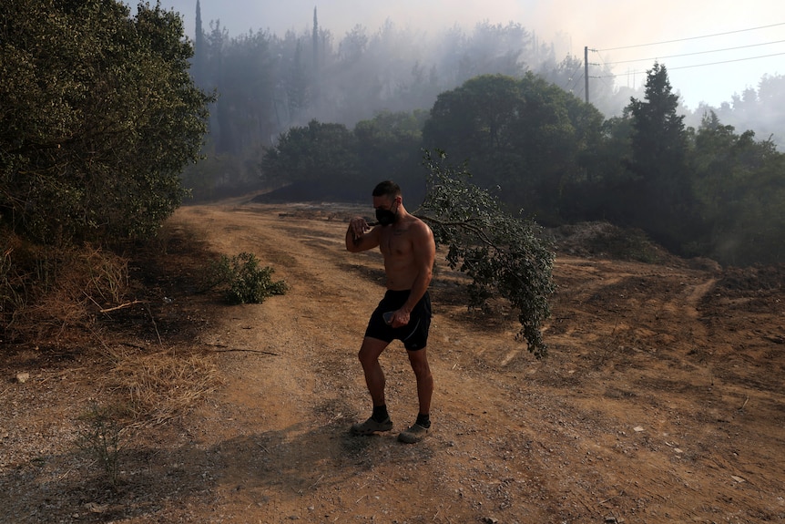 A local resident walks amidst smoke rising from a wildfire. He is wearing shorts with no top and a face mask.