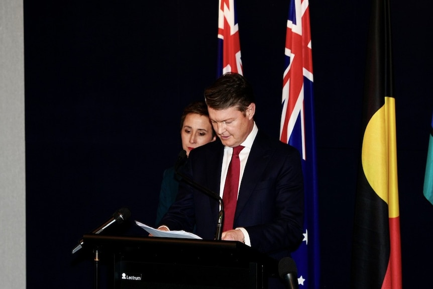 Ben Carroll speaks from a podium as Gabrielle Williams looks on