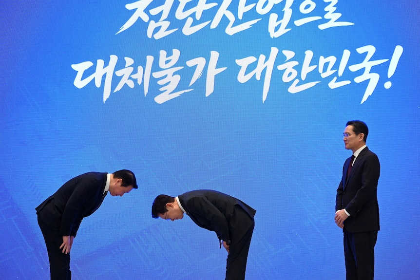 Two suited men bow deeply to each other on a stage as a third suited man watches on.
