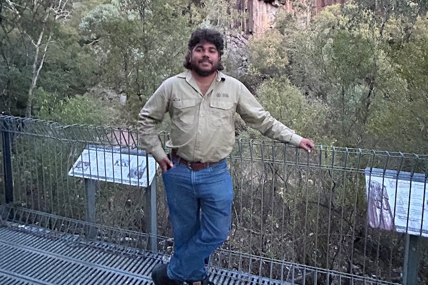 A smiling man leans on a safety rail, with trees and a tall, steep rock formation in the background.