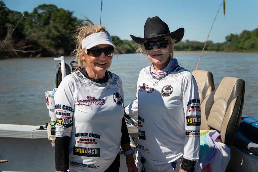 Two women in white longsleeve shirts, standing on a boat, with fishing gear around them, smiling at camera.