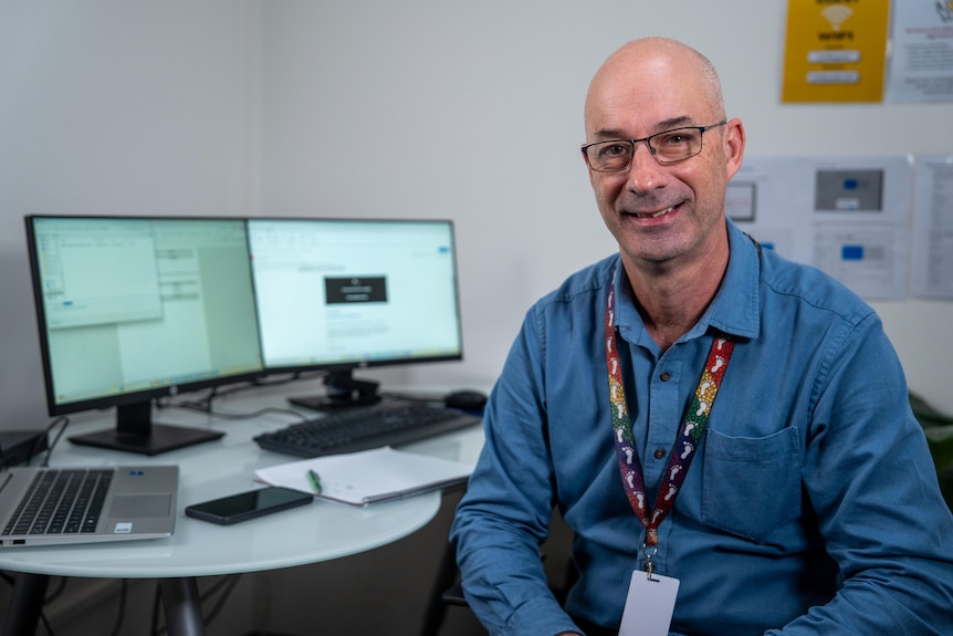 A man is bald and wears glasses and a blue shirt, he smiles while sitting in front of a computer