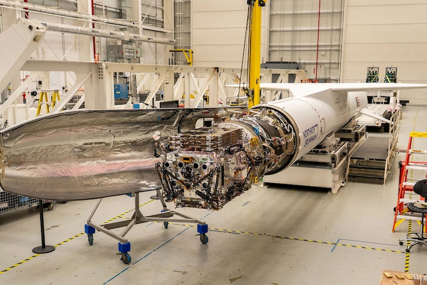 A small robotic space craft is attached to white rocket, and a carrying plane, inside a NASA facility.