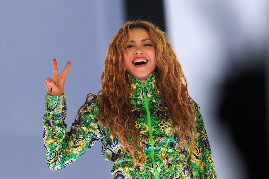 A woman with long hair and a green dress, smiles and makes the peace sign with her right hand.