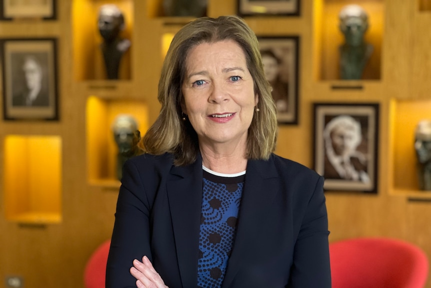 A smiling, middle-aged woman in a dark blazer stands in a wood-panelled room with portraits on the wall.