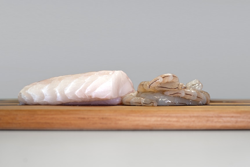 A piece of fish and a pile of shelled prawns on a wooden board.