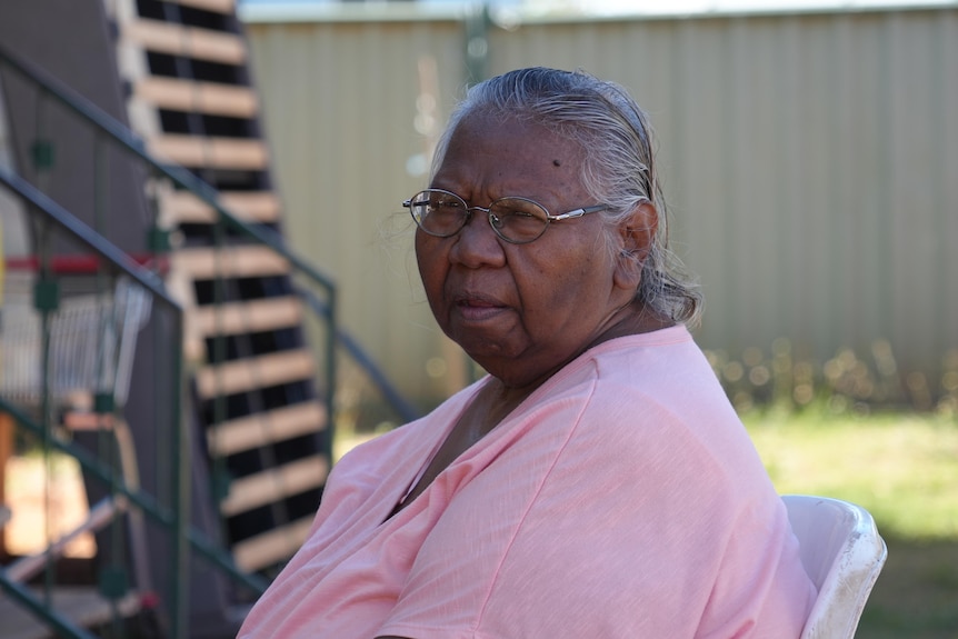 Christine looks to the camera over her left shoulder sitting on a white plastic chair with a pink shirt, grey hair and glasses