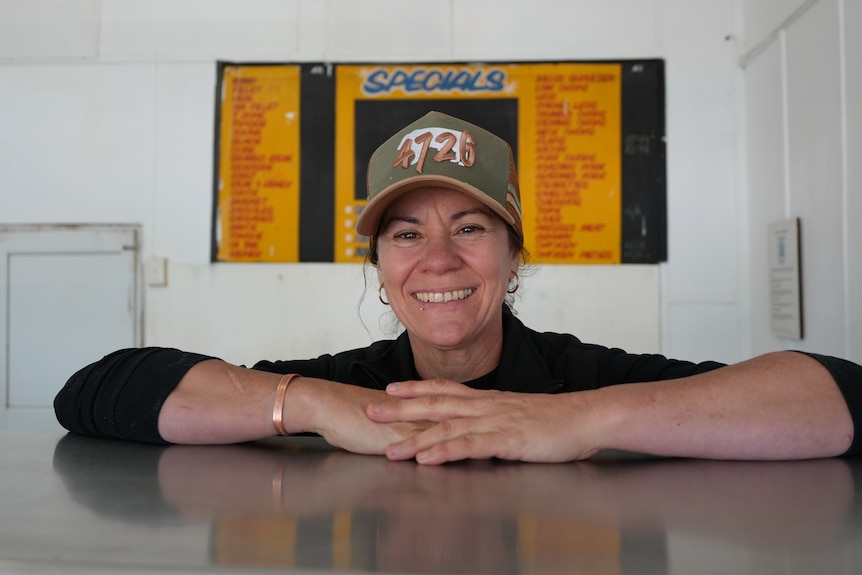 Woman standing behind counter at outback butcher shop smiling with her arms on the bench. She wears a black jumper and cap.