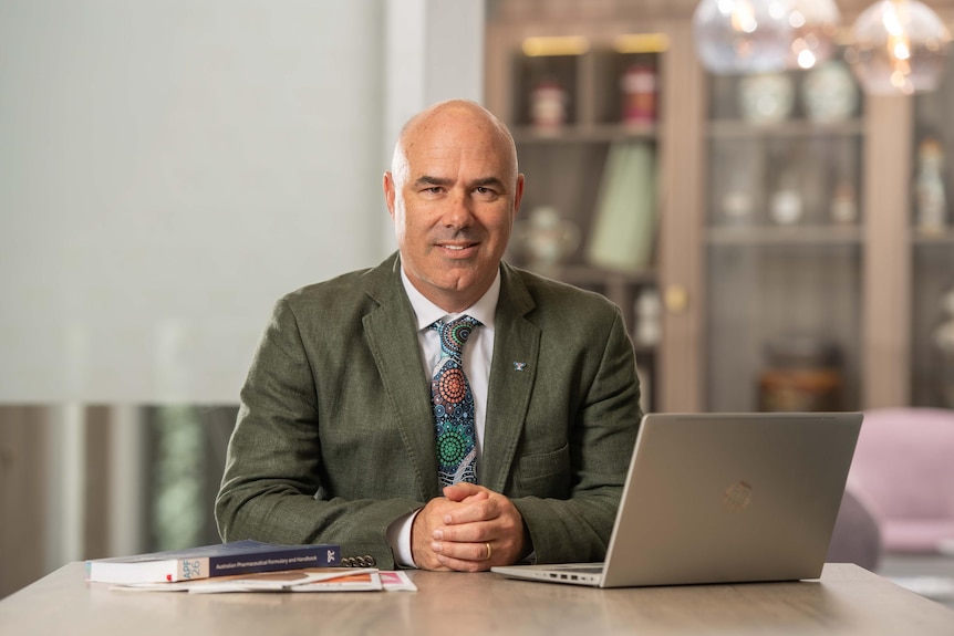 A man wearing a brown suit sits at a desk with books and an open laptop. He smiles at the camera.