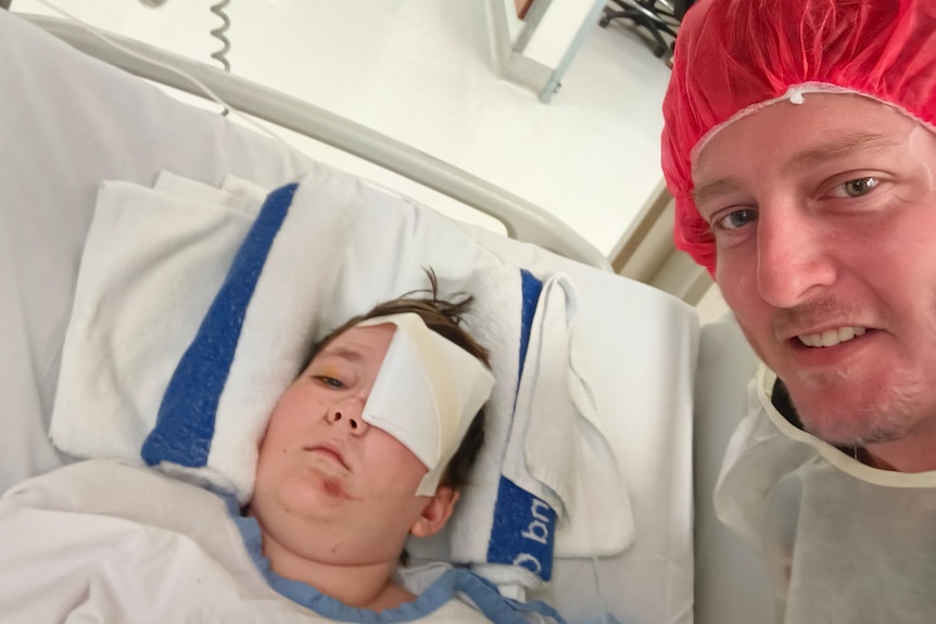 A boy with a patch over is eye lies in a hospital bed, his dad with a red medical cap and hospital gown takes a photo