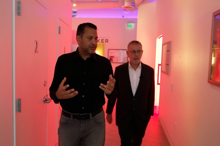 Darshan leads talking, gesturing with hands, while Norman follows, both in suits walking through a red-lit hallway in a clinic.