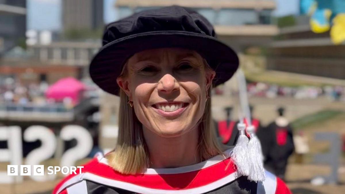 Beth Barrett-Wild in a doctoral-level academic dress, including a black Tudor bonnet hat. She is smiling at the camera and the background is blurred.